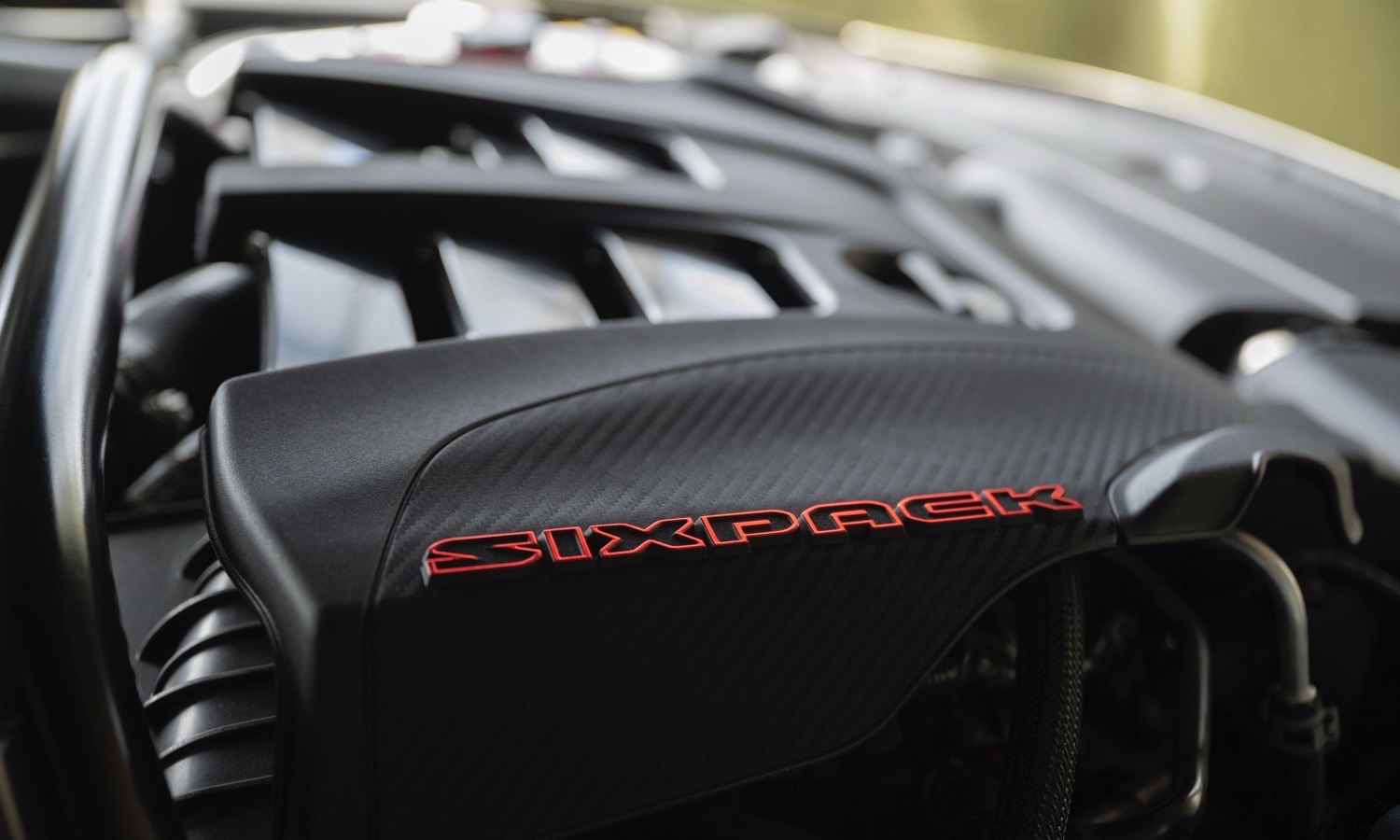 SIXPACK-powered Dodge Charger models feature a carbon-styled engine cover stamped with a red-accented SIXPACK text logo that announces the presence of the internal combustion engine under the hood.